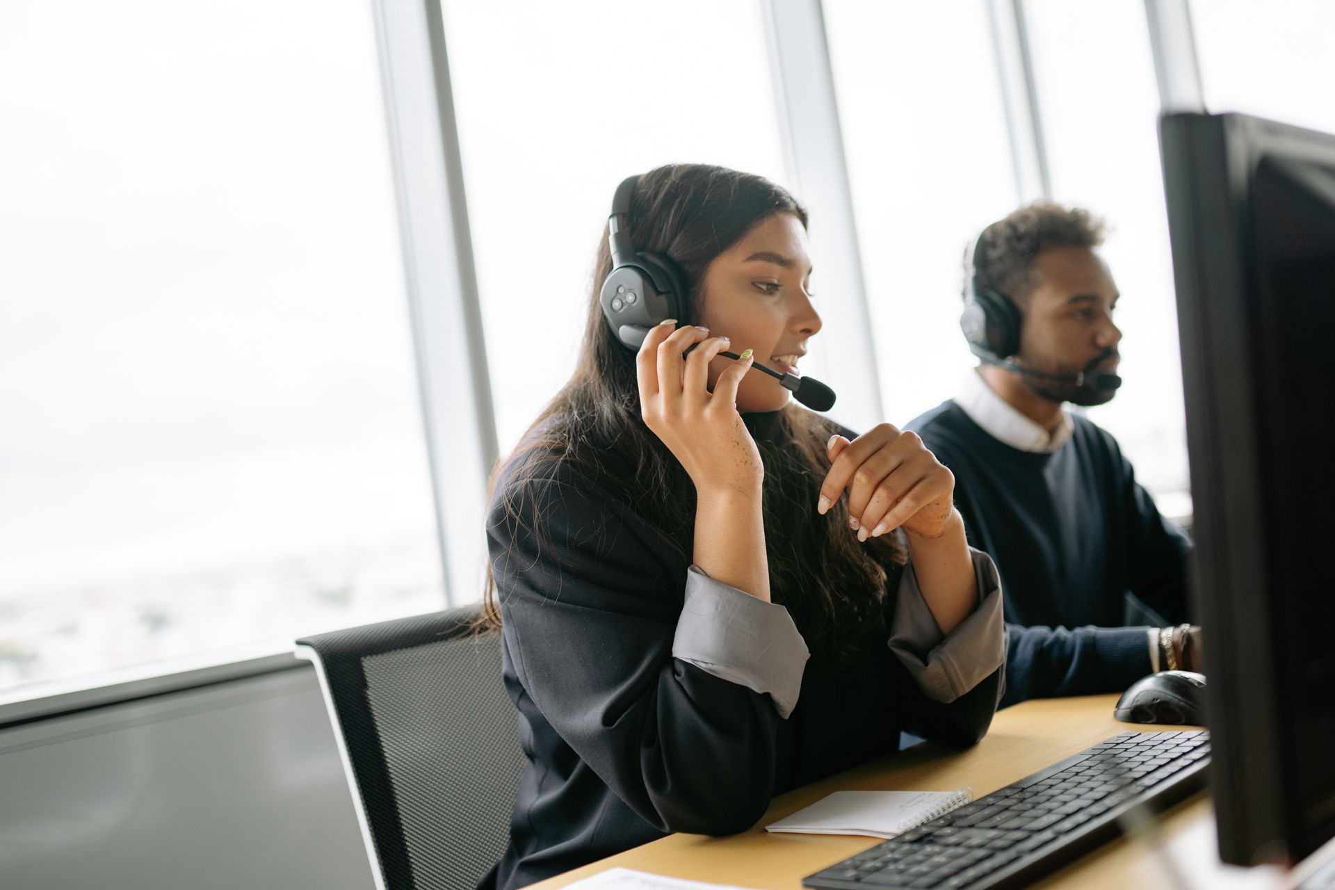 Two customer service agents wearing headsets answering calls at a shared desk in a bright office