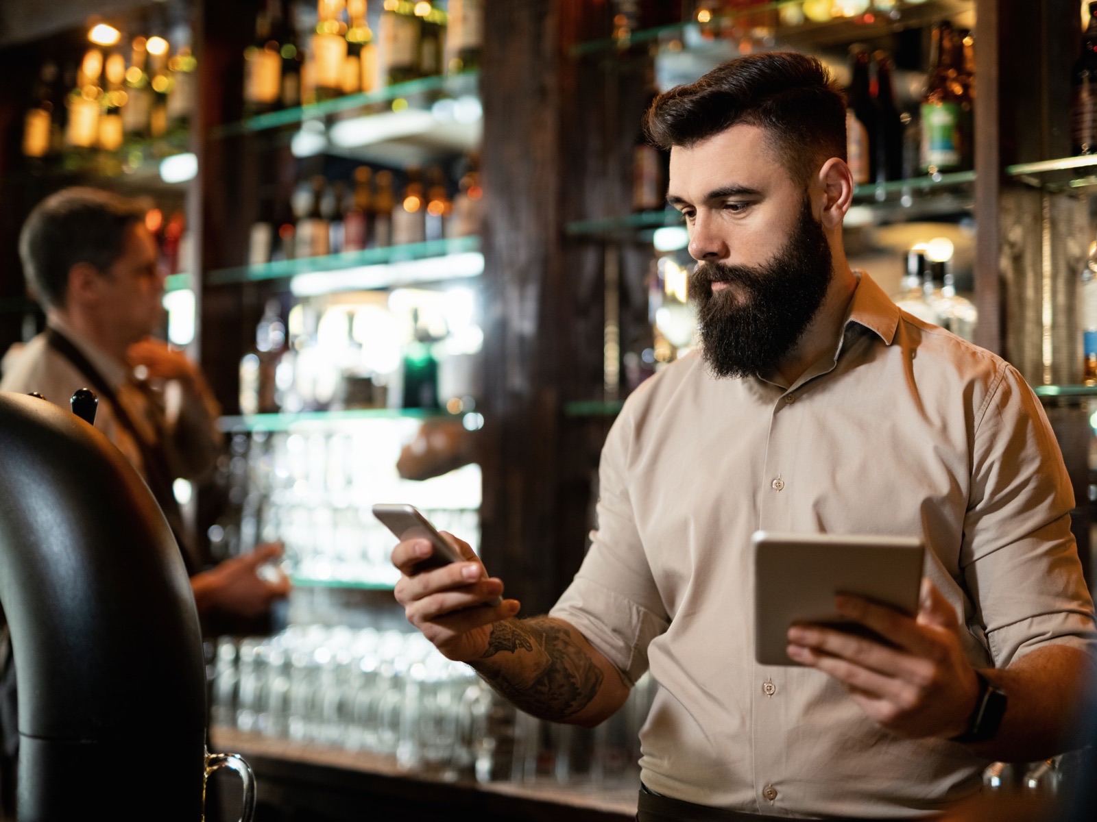 Bar owner answering customer messages on his phone while holding a tablet