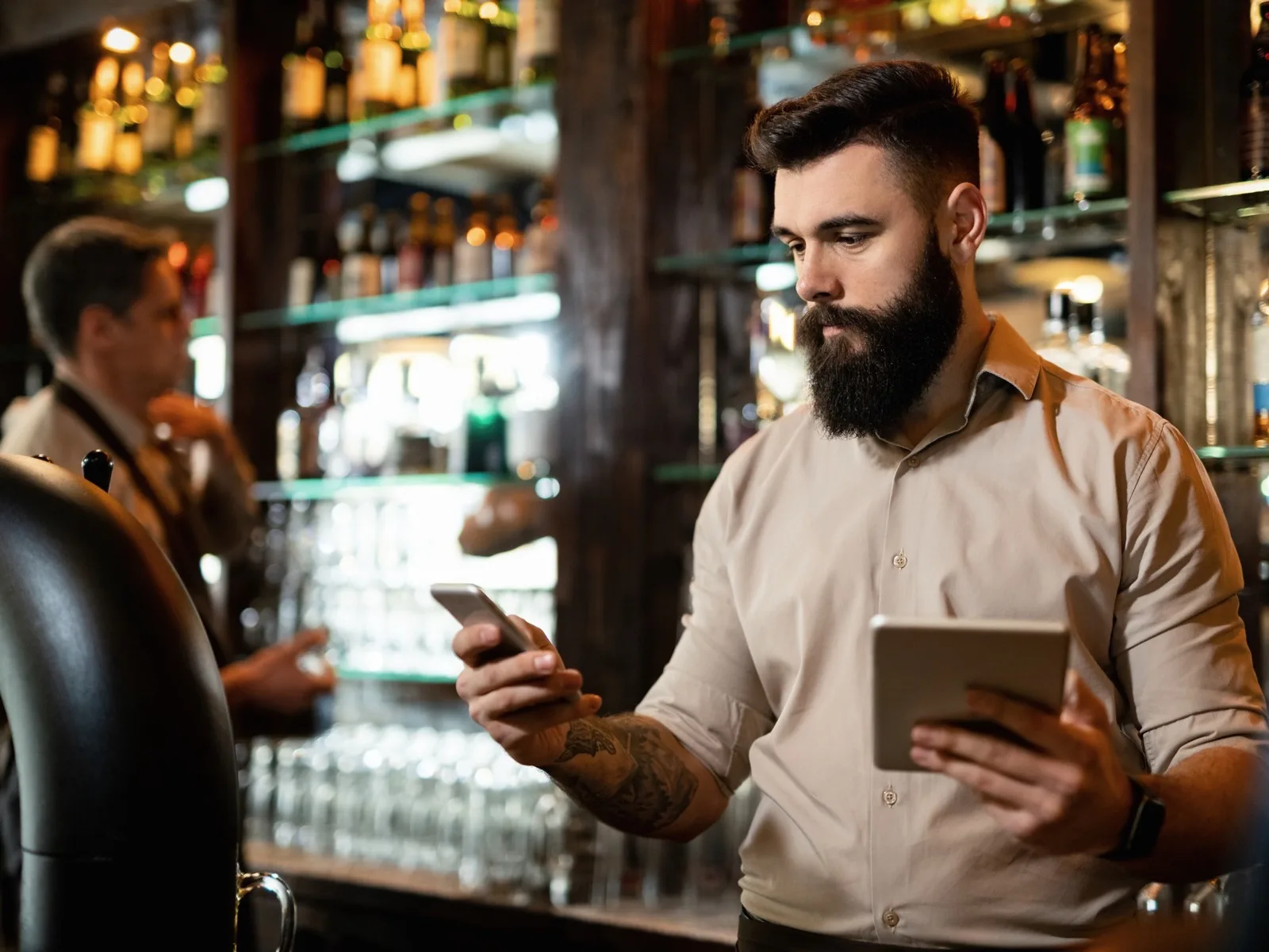 Bar owner answering customer messages on his phone while holding a tablet